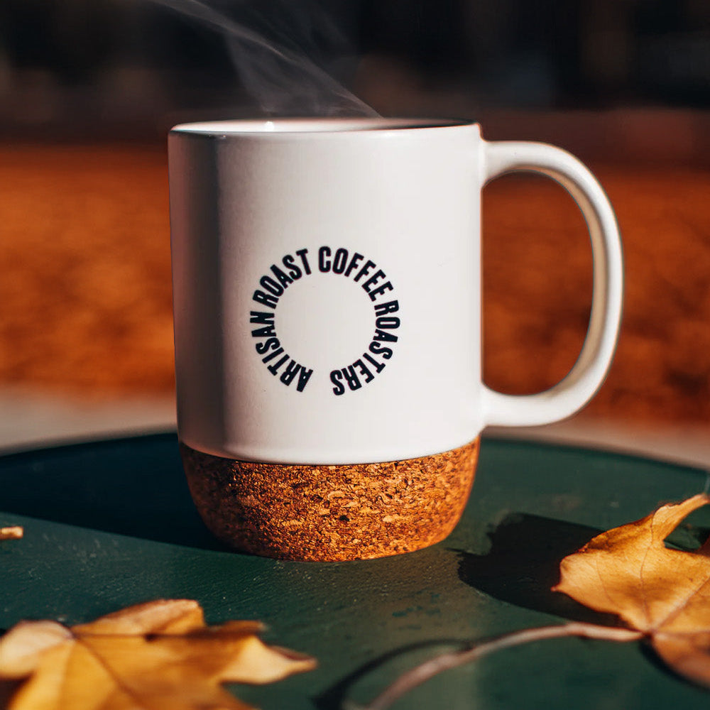 White mug with cork base on a green surface with autumn leaves, blurred background