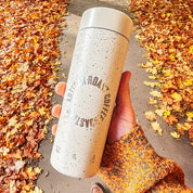 Person holding a speckled coffee cup with text on a path surrounded by autumn leaves