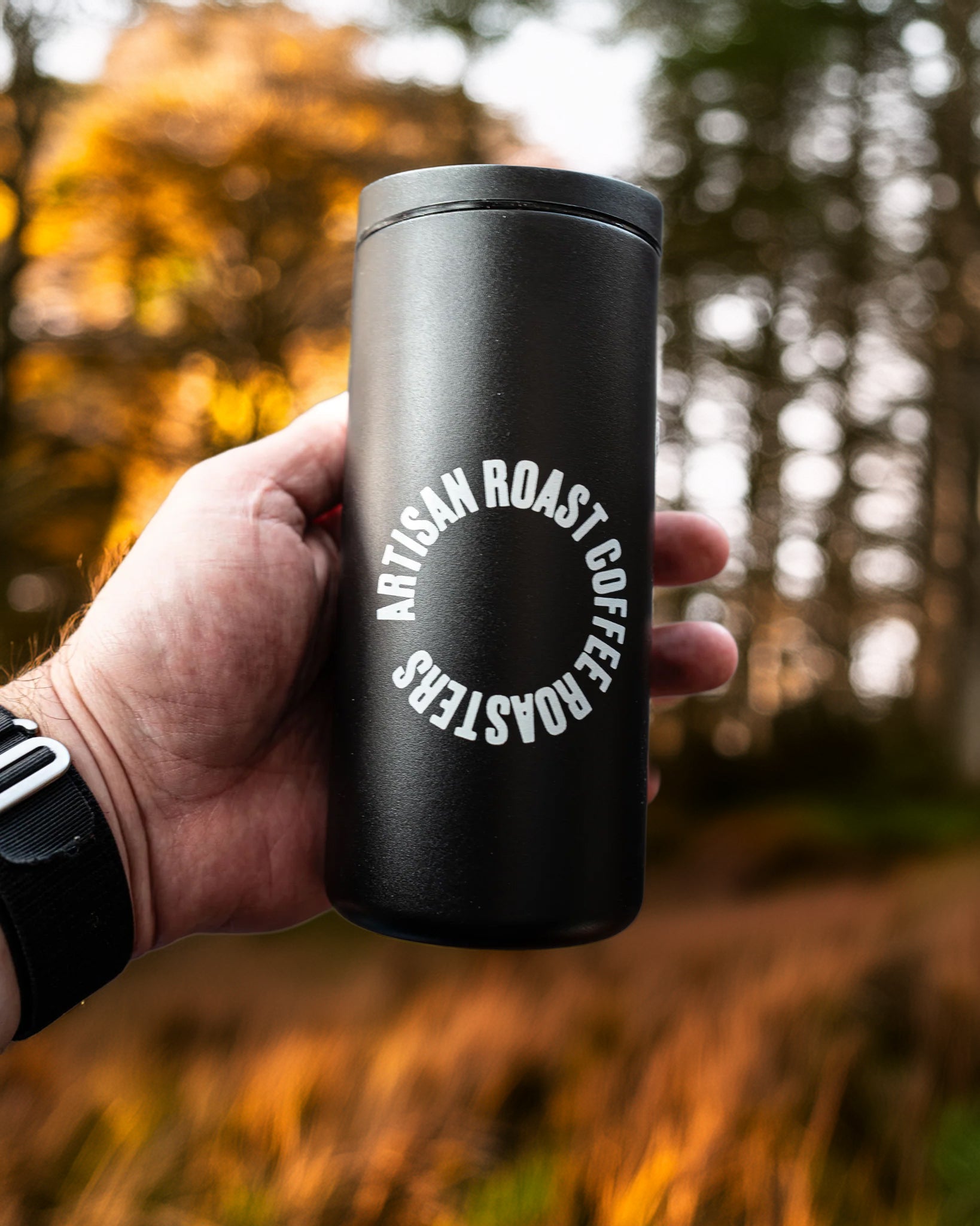 Black insulated coffee cup with 'Artisan Roast Coffee Roasters' logo held in a hand against a blurred forest background.